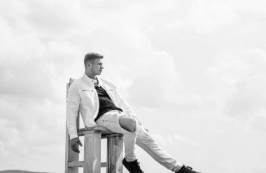 Gündelik tarzda giyinen yakışıklı genç adam tahta koltukta rahatla temiz havanın tadını çıkar gökyüzü bulutlu, fotokopi alanı için dinlen.