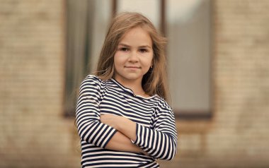 Özgüven içten gelir. Kendine güvenen bebek şehir dışında kollarını kavuşturuyor. Moda çocuğuna özgüvenli bir bakış. Modayı takip eden çocuklar giyer. Çocuk kıyafetleri. Sıradan moda trendi. Kendinden emin ve güzel.