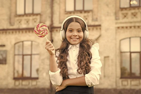 La ragazzina in uniforme mangia lecca-lecca. Torniamo a scuola. istruzione moderna con nuove tecnologie. Un ragazzo nel cortile della scuola. Immagina che sia una cantante. bambino ha pausa musica. ragazza in cuffia — Foto Stock