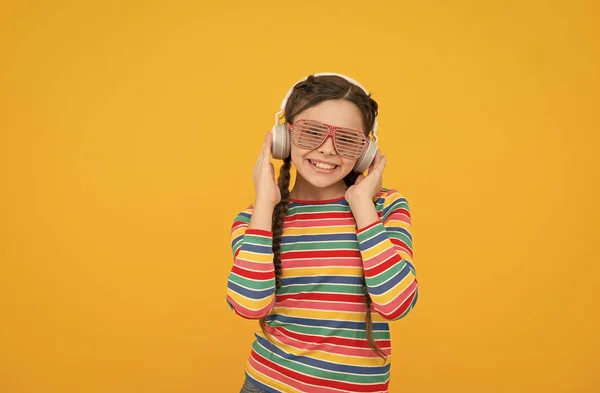 Veselé dítě dívka s copánky nosit barevné oblečení a okouzlující party brýle tanec a poslouchat hudbu ve sluchátkách na žlutém pozadí, melodie — Stock fotografie