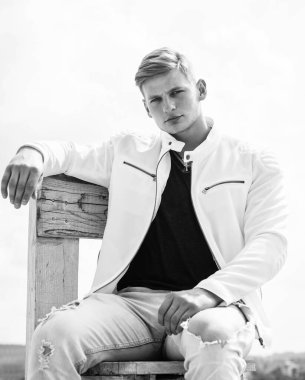 Yakışıklı, gündelik tarzda genç bir adam ahşap sandalyeye oturur açık havada bulutlu gökyüzü dalgın bir görünüm, ciddi.