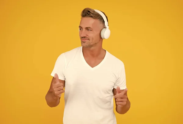 Homem de meia-idade feliz ouvir música em fones de ouvido modernos apontando dedos indicadores fundo amarelo, promovendo — Fotografia de Stock