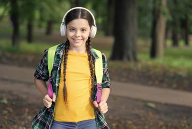 Mutlu çocuk modern kulaklıklarla müzik dersi dinliyor okul çantasını sıradan moda tarzında taşıyor doğal yaz manzarasını dinliyor.