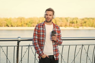 Kahve fincanı açık havada traşsız bir adam. Gündelik erkek hippi modası. Kulaklıklı adam, dinle. Müzik tarzı. Kahve içerken para kazanmak. Ses kalitesi. Günaydın içecek.