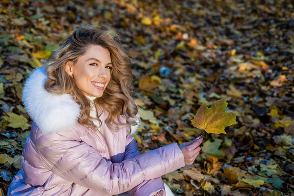 Gelukkig jong vrouw met herfst look glimlach zitten op gevallen bladeren in warm outdoor kleding, meisje — Stockfoto