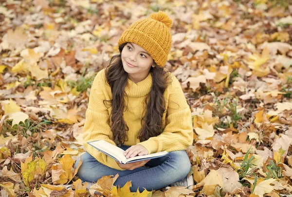 Pequena criança ler livro com olhar pensativo sentado em folhas de outono ao ar livre, imaginação — Fotografia de Stock