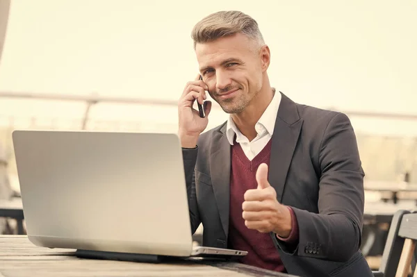幸せなビジネスマンは、携帯電話でクライアントと話をし、オンラインビジネスのためのコンピュータを使用して手のジェスチャーを親指を表示,承認 — ストック写真
