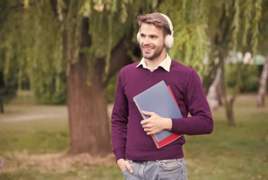 Modern kolej öğrencisi parkta durmuş kulaklıkla müzik dinliyor ve kitap okuyor.