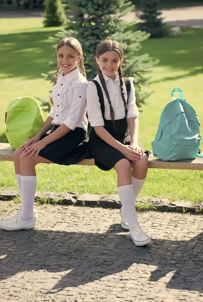 Niños felices en uniforme formal se sientan en el banco del parque con bolsas relajantes después del día escolar al aire libre, los alumnos — Foto de Stock