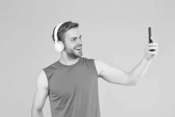 Χαρούμενος νεαρός άνδρας στα αθλητικά ρούχα φορούν μοντέρνα ακουστικά ακούγοντας μουσική, ενώ η κατάρτιση χρησιμοποιούν smartphone για selfie, ψηφιακό αθλητισμό — Φωτογραφία Αρχείου