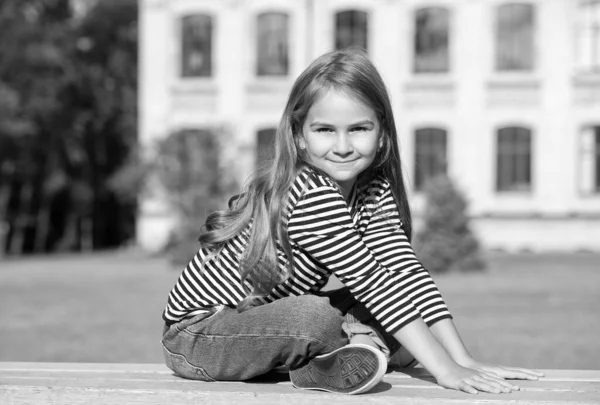 Счастливый маленький ребенок с длинными волосами в стиле casual сидит на скамейке парка, расслабляясь в солнечный летний день на открытом воздухе, отдыхая — стоковое фото