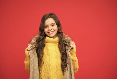 Çocuk sonbahar moda tarzında. Mutlu çocuk örgü süveter ve yelek giyer. Genç kızın uzun kıvırcık saçları var. Mutlu çocukluk. Kırmızı arka planda modaya uygun bir çocuk. Kalın giysiler ve örgü.