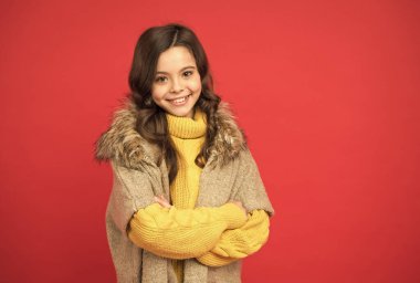 Gülümseyen tatlı çocuk uzun kıvırcık saçlı kalın örgü süveter giyiyor ve kırmızı arka planda sonbahar yeleği giyiyor, sezon tarzı.