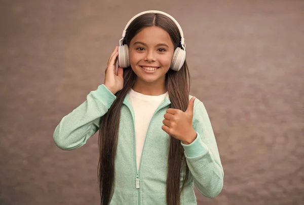Szczęśliwe uśmiechnięte dziecko lubi słuchać muzyki w słuchawkach. Dzieciak słucha piosenki na zewnątrz. Chodzenie z ulubioną melodią. edukacja online koncepcja. szczęście z dzieciństwa. elearning uczennicy. kciuk w górę — Zdjęcie stockowe