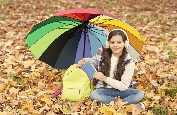 Vidám tini gyerek pihenni őszi erdőben vagy parkban, miközben zenét hallgatni headset könyv az iskolai hátizsák alatt színes esernyő, e-learning — Stock Fotó