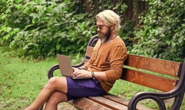 Sakallı adam dizüstü bilgisayarla bankta oturuyor. Online uzak eğitim. Hipster parkta çalışmak için ilham aldı. Çevik iş. Bilgisayarda çalışan olgun bir öğrenci. Pazartesi günü alışverişe git.