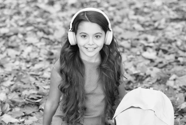 Happy childhood. kid listen music in autumn park. fall is a time for study. back to school. concept of online education. child sit in autumn leaves. girl in earphones with school bag — Stock Photo, Image