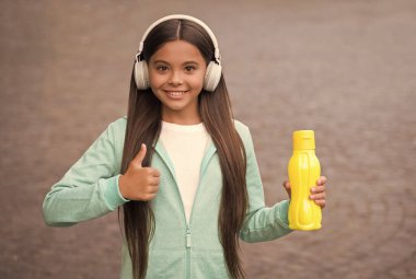 Çocuk vücudunda su dengesi. Öğle yemeği zamanı. Sıvı kaybı. Sağlıklı bir yaşam tarzı. çocukluk mutluluğu. Mutlu çocuk dışarıda kulaklık takıyor. Okula dönüyorum. Genç kız spor şişesini tutuyor. başparmak hareketi