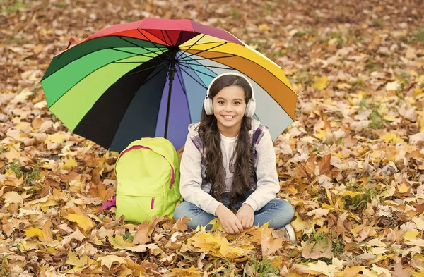 Anak bahagia mendengarkan musik di headphone sambil duduk di hutan musim gugur di antara daun jatuh dengan payung berwarna-warni dan ransel sekolah, kembali ke sekolah — Stok Foto