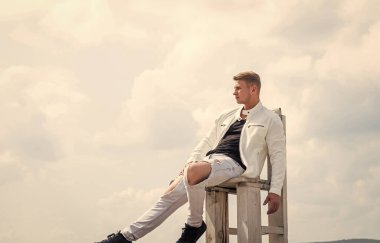 Gündelik tarzda giyinen yakışıklı genç adam tahta koltukta rahatla temiz havanın tadını çıkar gökyüzü bulutlu, fotokopi alanı için dinlen.