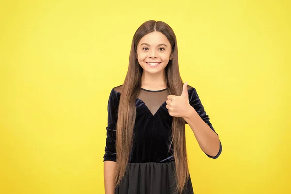 Criança menina feliz com cabelos longos morena e beleza olhar desgaste vestido de moda mostrando polegares ups mão gesto amarelo fundo, aprovação — Fotografia de Stock