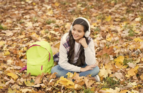 Boldog kisgyerek zenét hallgat fülhallgatóban, őszi leveleken ül a szabadban, vissza az iskolába. — Stock Fotó