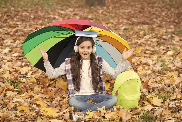 Jesienny dzieciak pod parasolem. Przeczytaj książkę. Szczęśliwego dzieciństwa. Z powrotem do szkoły. dziewczyna w słuchawkach z plecakiem zrelaksować się w parku. cieszyć się jesienią w lesie. Słuchaj muzyki. edukacja online — Zdjęcie stockowe