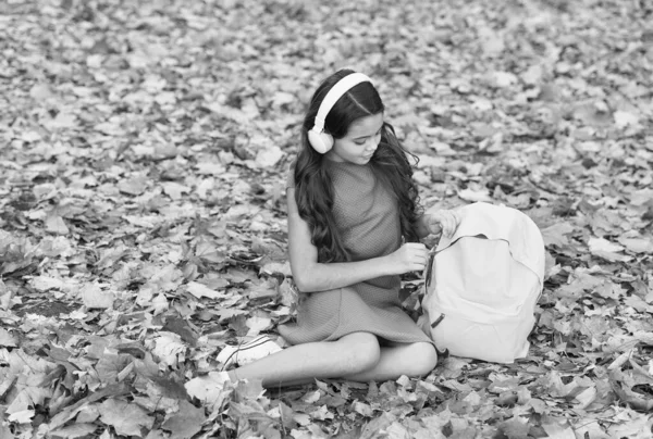학교 가방을 들고 다니는 소녀. 아이는 가을 파크에서 음악을 듣습니다. 가을은 연구 할 때입니다. 다시 학교로. 온라인 교육 개념. 가을 나뭇잎에 앉아 있는 아이 — 스톡 사진