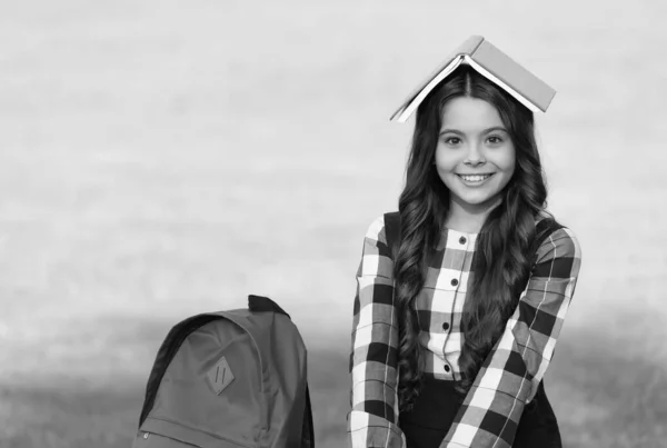 Joyeux petit enfant en uniforme scolaire tenir livre d'étude sur la tête sur le paysage naturel, fantaisie — Photo