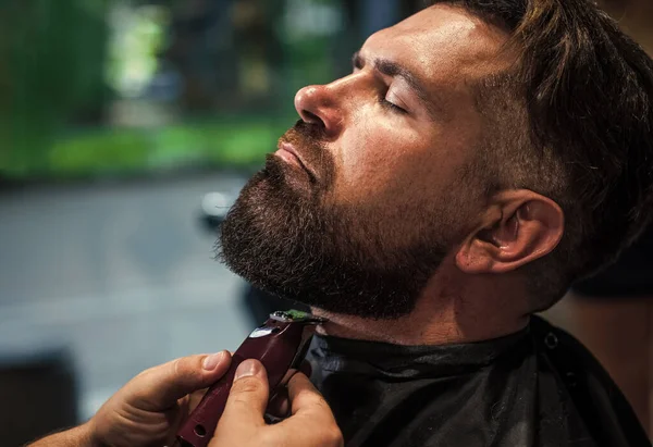 Capelli e barba styling. brutale ragazzo con maestro barbiere uso rasoio elettrico. hipster. cura barbiere maschile. L'uomo barbuto dal parrucchiere. bellezza e moda. Toccando la sua barba perfetta — Foto Stock