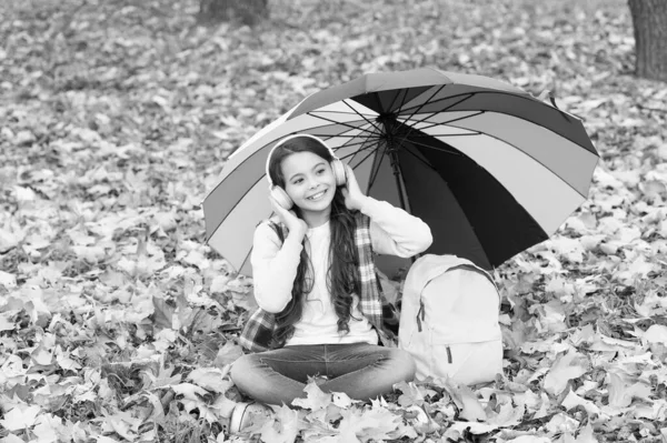 Neşeli genç kız okul çantasının yanında renkli şemsiyeler altında kulaklıkla müzik dinlerken sonbahar ormanlarında ya da parkta dinleniyor. — Stok fotoğraf