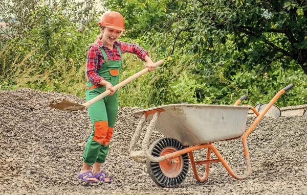 Девочка-строитель в защитном шлеме использовать лопату и строительную тачку, строительную площадку — стоковое фото