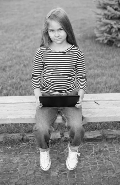 Adorable small kid in casual style with smart look use modern tablet sitting on park bench outdoors, computer — Stock Photo, Image