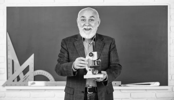 Professor sênior feliz usar microscópio. química no laboratório da escola. experimentos científicos com microscópio em laboratório. Biologia equipamentos de laboratório da escola. homem em sala de aula com ferramentas de matemática no quadro negro — Fotografia de Stock