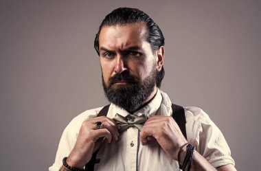 Bıyıklı vahşi sakallı adam pantolon askısı ve papyon takıyor, modası geçmiş gibi görünüyor.