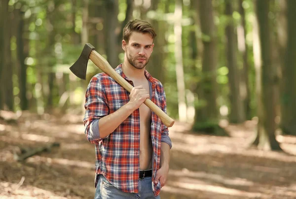 Unshaven homem bonito em camisa xadrez aberto com jeans transportar grande machado rachando na floresta de verão paisagem natural, lenhador — Fotografia de Stock