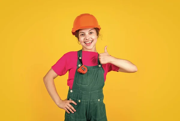 Kid wear helmet on construction site. teen girl builder with building tool tape-measure. child on repairing work. concept of renovation in workshop. busy professional carpenter show thumb up — Stock Photo, Image