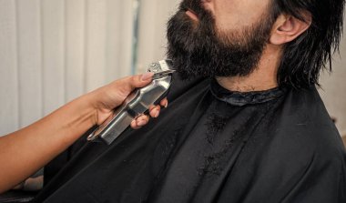 Güzellik ve moda. Saç ve sakal şekillendirme. Profesyonel berber ustası olan kendine güveni tam acımasız bir adam. Hippi için elektrikli tıraş makinesi. Erkek berber bakımı. Kuaför salonundaki sakallı adam.