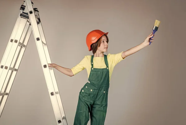 Веселая девушка-подросток в рабочей форме и шлеме на лестнице использовать кисть, художник — стоковое фото
