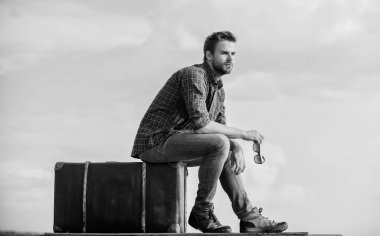 Macera orada bir yerde. Seksi erkek gökyüzü arka planı. Erkek modası. Çok modaya uygun görünüyorsun. Gözlüklü iş adamı. İş gezisi. Maço, tıraşsız bir adam tur çantasına oturur. Gezgin, uçuşu bekle. Taşı