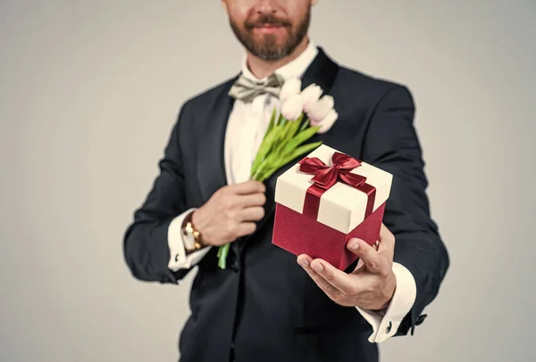 Бородатий чоловік обрізаний вид тримає подарункову коробку, пов'язану з червоною стрічкою лук вибірковий фокус і весняні квіти для святкування сірого фону, 8 березня — стокове фото