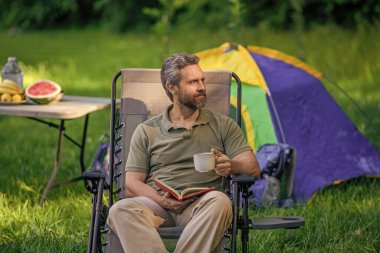 Adam ormanda sabah kampı kahvesi içiyor. Yürüyüşçü adam dışarıda rahatlar. Yaz kampı ve doğa tatili. Adam kamp hakkında kitap okudu. Kamp macerası. Yürüyüş konsepti. Kamp çadırı. Kahve ve kitap..