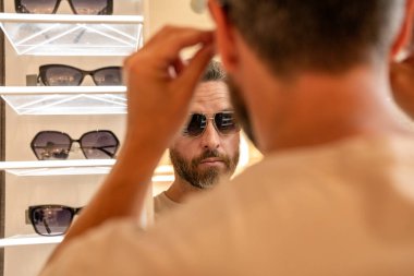 Dükkanda güneş gözlüğü seçen bir adam. Moda mağazasında güneş gözlüğü mankeni. Modaya uygun bir adam. Erkekler için moda güneş gözlüğü aksesuarları. Göz giysisi koleksiyonu. Güneş gözlüklü, seçici odaklı moda görünümü.