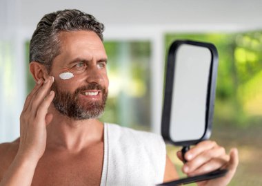 Deri kozmetiği. Güzellik kremi ya da yaşlanmayı önleyen cilt bakımı. Erkekler için makyaj malzemesi. Erkek yüzü için krem. Yaşlanma önleyici ve kırışıklık önleyici krem. Adam aynaya kozmetik yüz kremi sürüyor..