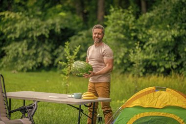 Kamp ve doğa pikniği. Adam kampı. Karpuz kampı pikniği. Lezzetli sağlıklı yiyecekler. Kampta taze yiyecek. Yaz kampında elinde karpuz tutan adam. Yaz günü erkek yürüyüşçüsü. Meyve yiyin..