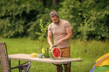 İspanyol olgun bir adam yaz kampı pikniğinde yemek için karpuz kesti. Lezzetli ve sağlıklı..