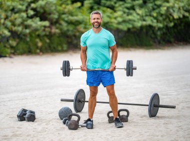 İspanyol adam halterle dışarıda egzersiz yapıyor. Erkek sporcu halter kaldırıyor. Güçlü adam spor egzersizi yapar. Jimnastik. Halterli sporcu. Sağlıklı yaşam halter eğitimi.