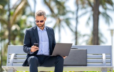 İş adamı parkta dizüstü bilgisayarla sohbet ediyor. İnternette çalışan bir iş adamı. Başarılı iş adamı çevrimiçi olarak serbest çalışıyor. İş başarısı. İş adamı dışarıda dizüstü bilgisayarda çalışıyor..