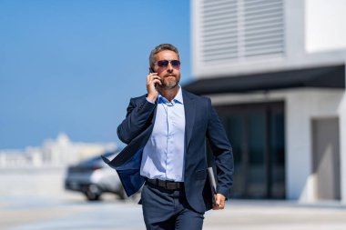 Başarılı bir iş adamı otoparkta dizüstü bilgisayarla yürüyor. Takım elbiseli olgun bir iş adamı iş görüşmesi yapıyor. Şirket işi. Takım elbiseli zengin iş adamı. İş Portresi.