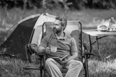 Adam ormanda sabah kampı kahvesi içiyor. Yürüyüşçü adam dışarıda rahatlar. Yaz kampı ve doğa tatili. Adam kamp hakkında kitap okudu. Kamp macerası. Yürüyüş konsepti. Kamp çadırı. Kahve ve kitap..